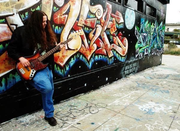 The site's author holding a bass guitar in front of a wall covered in graffiti